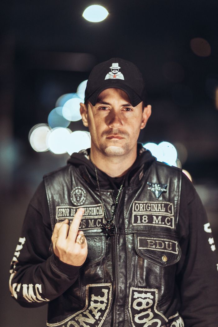 A man at night wearing a leather jacket and hat, making a defiant hand gesture.