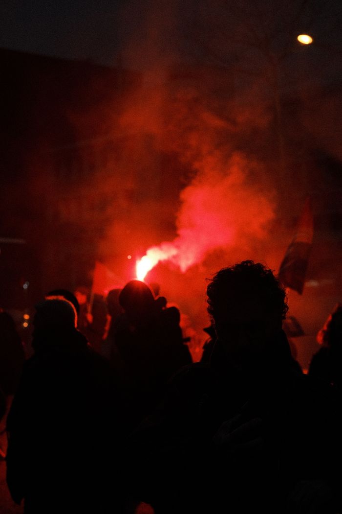 Silhouetted protest crowd with a red flare creating smoke at night, evoking strong emotions.