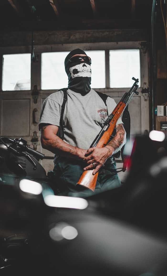 A masked person stands in a garage holding a rifle, surrounded by motorcycles.