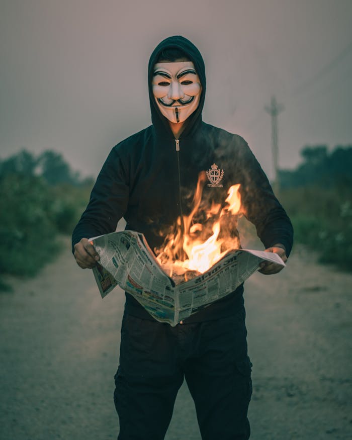 A person in a Guy Fawkes mask holds a burning newspaper, evoking mystery and rebellion.