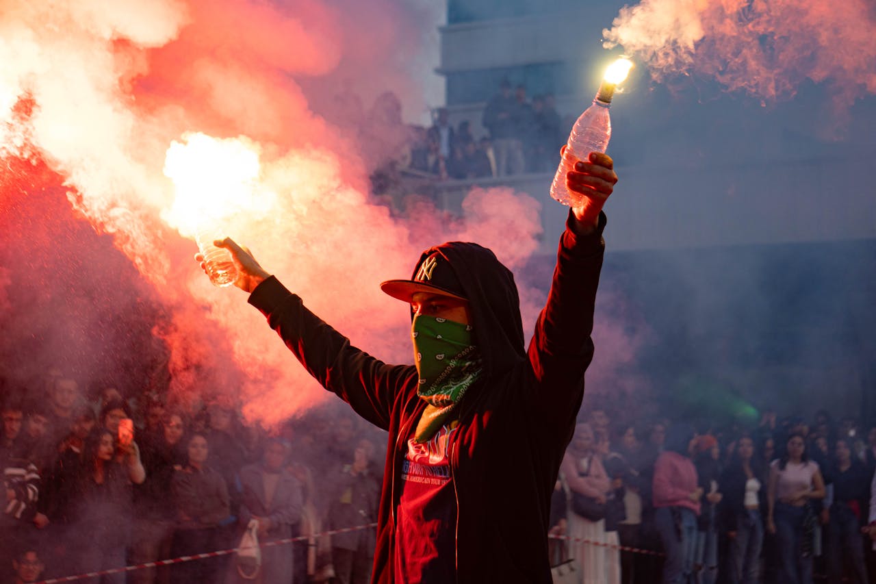 Protestor raising smoke flares amidst a large crowd in an outdoor urban setting.