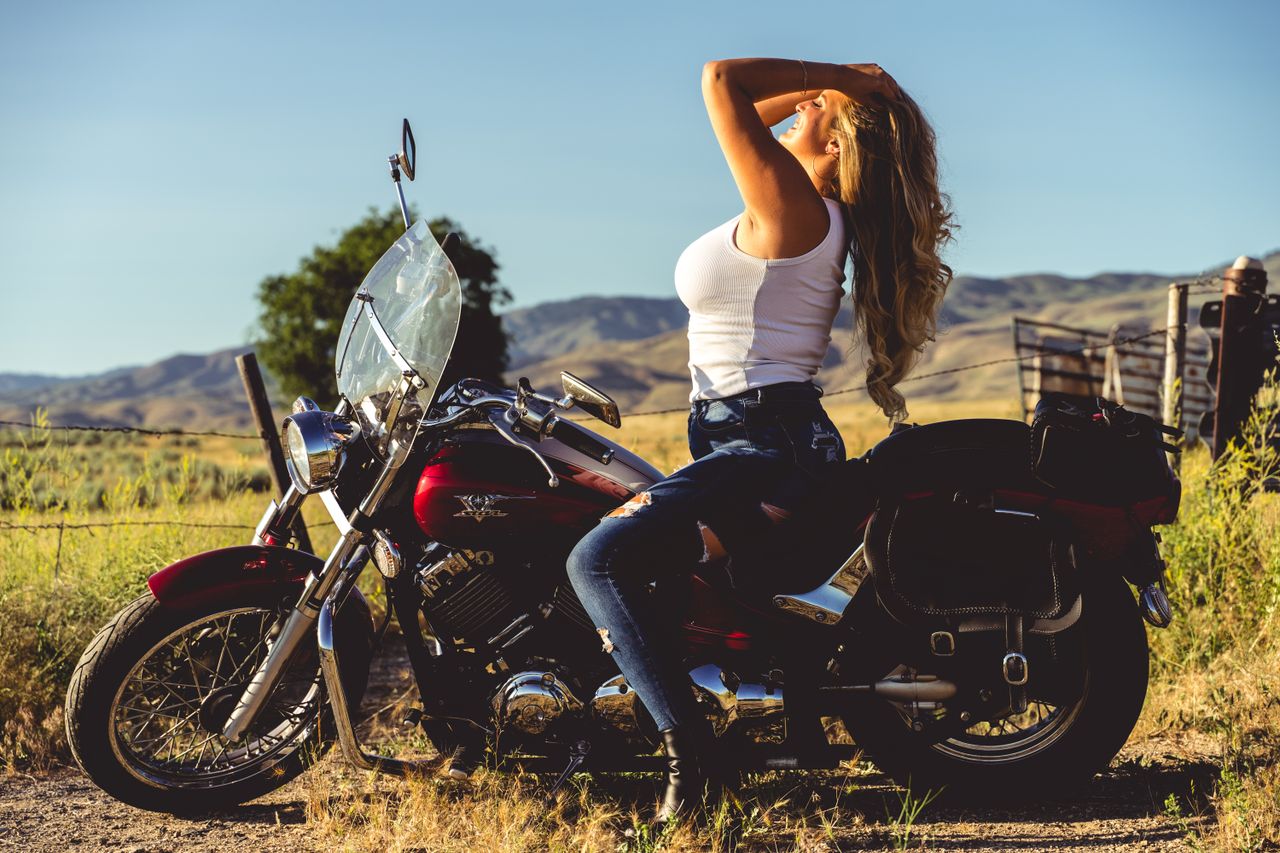 A woman in jeans and a tank top poses on a motorcycle in a scenic countryside.