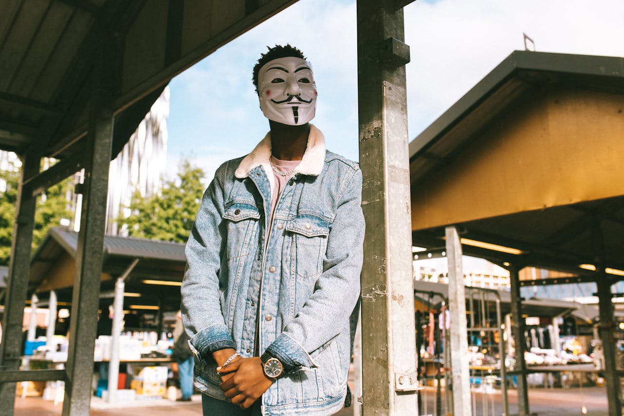 From below of worried African American activist in stylish clothes and famous Anonymous mask standing on street near local market in city and looking away in daytime